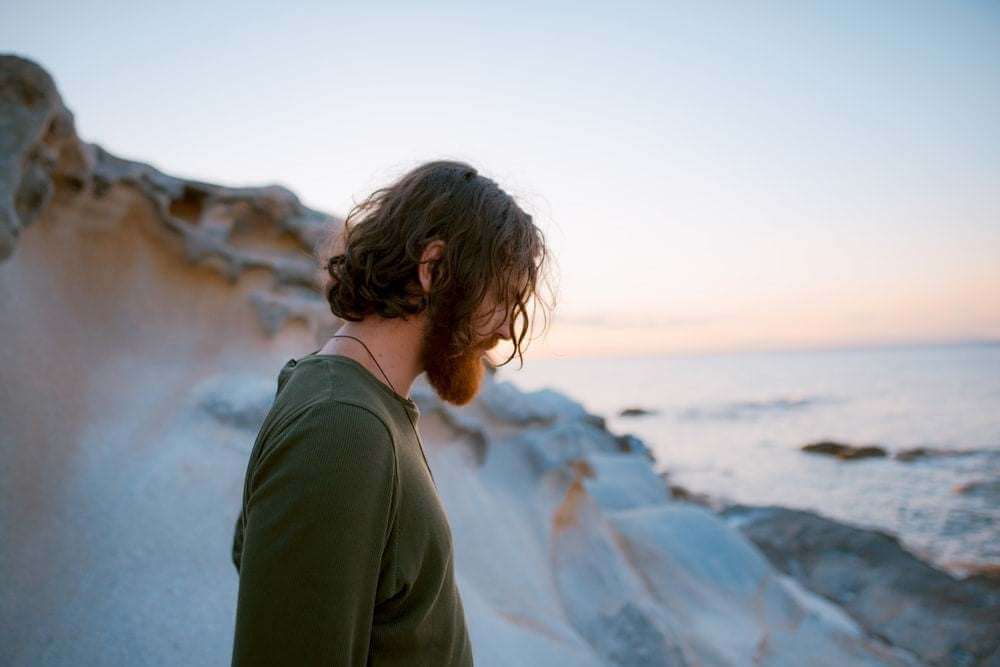man with a beard standing by rocky shore at sunset focusing on beard grooming with folding beard comb